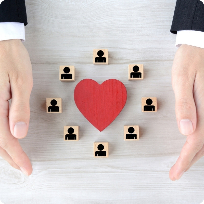 Two hands reaching towards a red heart with small wooden blocks representing people on a light gray background.