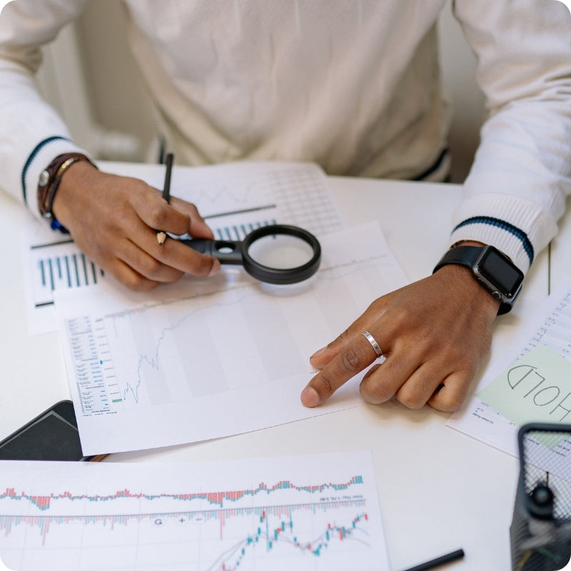 Person analyzing financial documents with a magnifying glass on a desk.