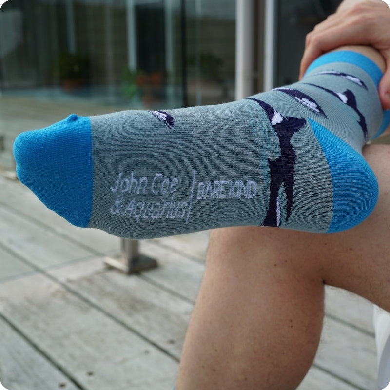 Person wearing a sock with 'John Coe & Aquarius' and 'Barekind' branding on a wooden deck.