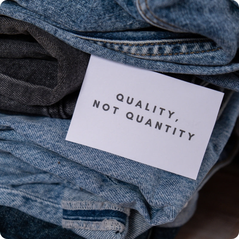 A close-up of folded blue denim jeans with a white paper tag printed with the text 'QUALITY, NOT QUANTITY' in black sans-serif lettering.