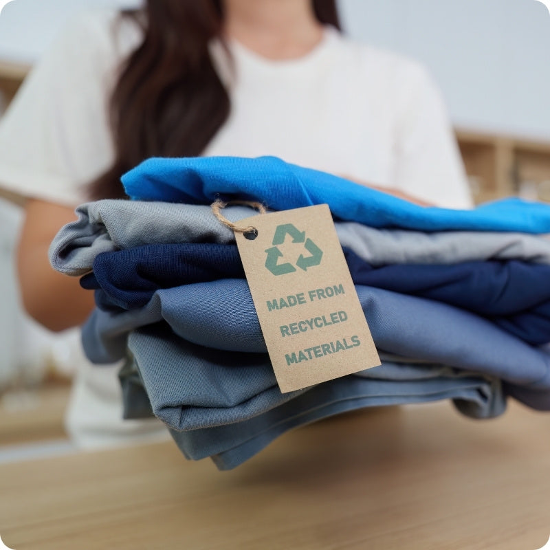 A stack of folded blue and gray garments held by a person in a white shirt, with a kraft paper tag reading 'MADE FROM RECYCLED MATERIALS' attached. The background is softly blurred, showing an indoor setting.