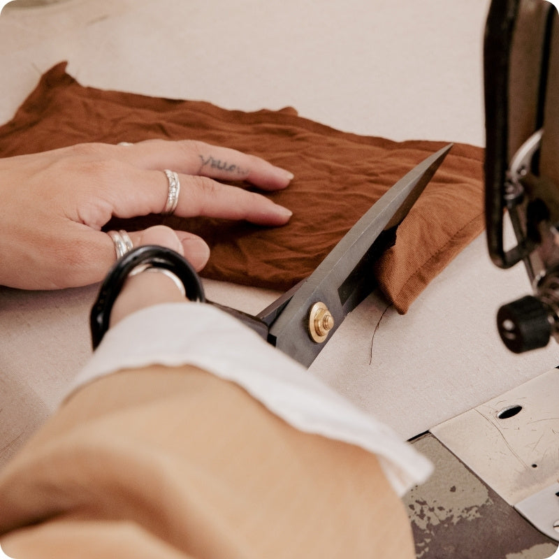 A close-up of a person's hands using large black-handled scissors to cut a brown fabric piece on a sewing machine table.