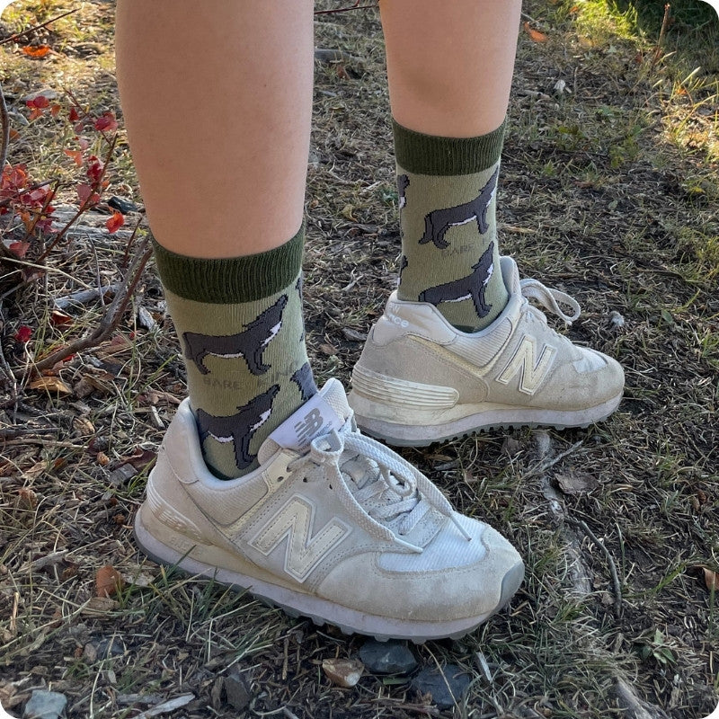 A person wearing olive green crew socks with a black wolf  pattern, paired with off-white New Balance sneakers, standing outdoors on a grassy, dirt-covered ground.