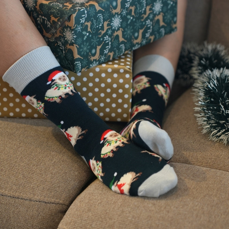 A person sitting on a beige couch wearing black Christmas socks with a sheep pattern, next to a gold and white polka dot gift box and holiday tinsel.
