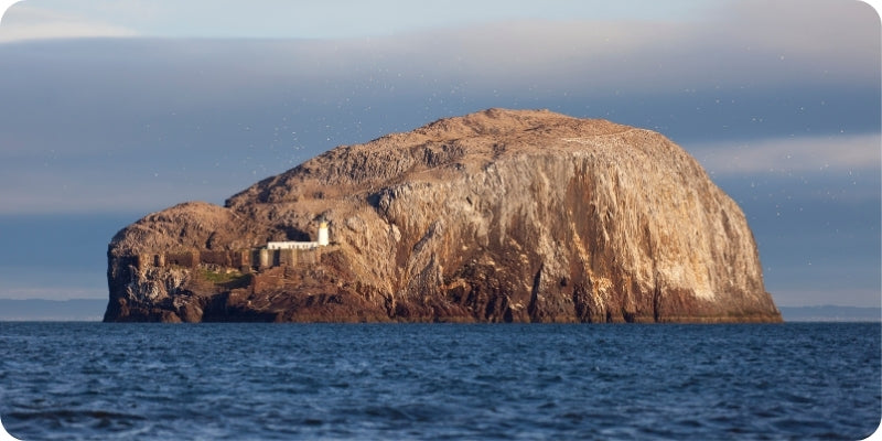 A large rocky island or headland rising from blue water under a cloudy sky, with a small white building near the base of the rock.