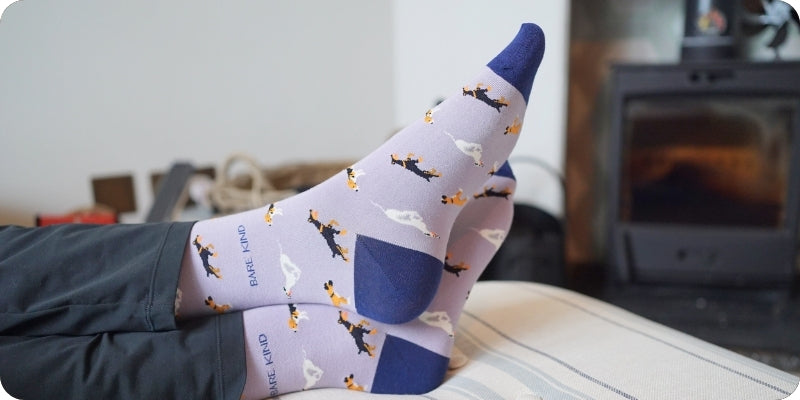 A person's feet wearing light purple socks with a pattern of small dogs and white clouds, resting on a bed in a cozy room with a fireplace in the background.
