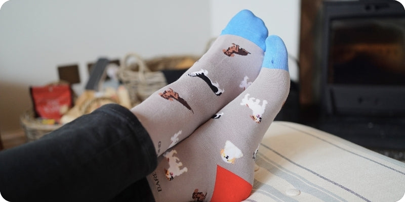A person's feet wearing gray socks with a pattern of small dogs and boots, blue toes, and orange heels, resting on a striped cushion in a cozy indoor setting.