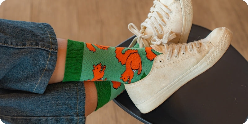 A person wearing green crew socks with an orange cartoon dog pattern, paired with blue jeans and off-white canvas sneakers, sitting on a black stool.
