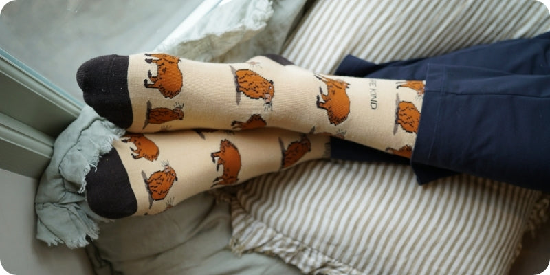 A person's legs crossed while wearing cream-colored socks with a brown capybara pattern, paired with navy blue pants, resting on a striped pillow.