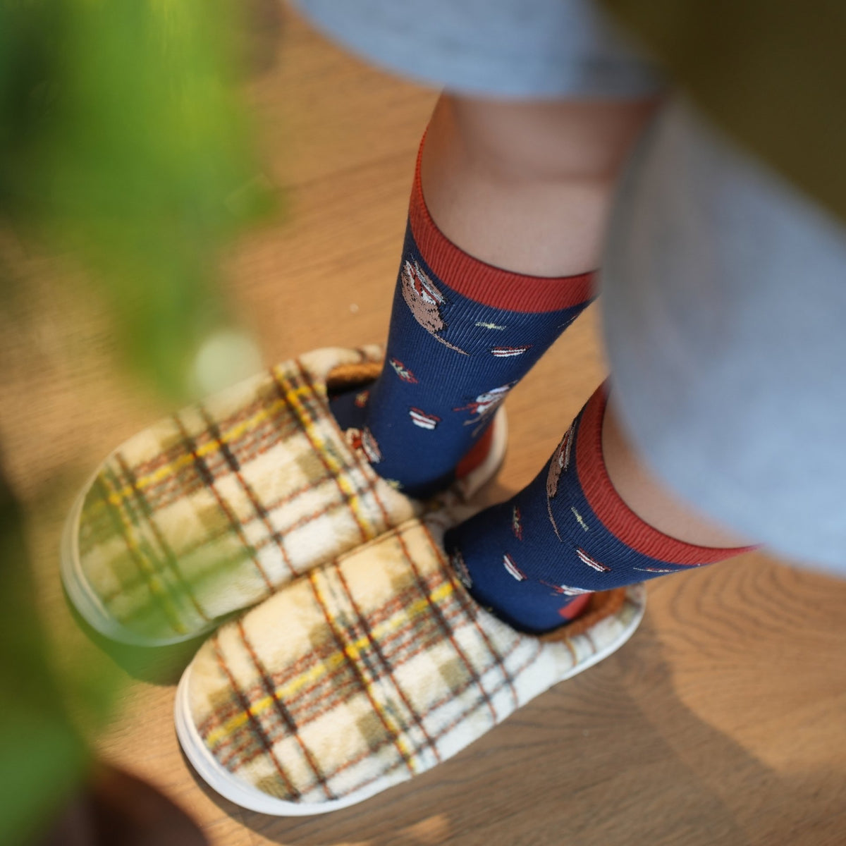 A close-up lifestyle shot of a person's feet wearing navy blue otter socks with a red cuff and a small pattern, paired with cream-colored slippers, standing on a wooden floor.