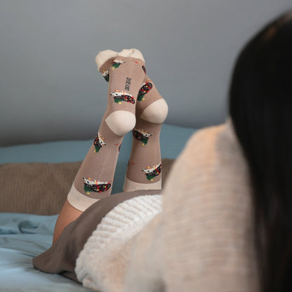 A person reclining on a bed with their legs raised, wearing beige knee-high socks with colorful cartoon hedgehogs prints and white heels. The background shows a gray wall and a blue-gray bedspread.