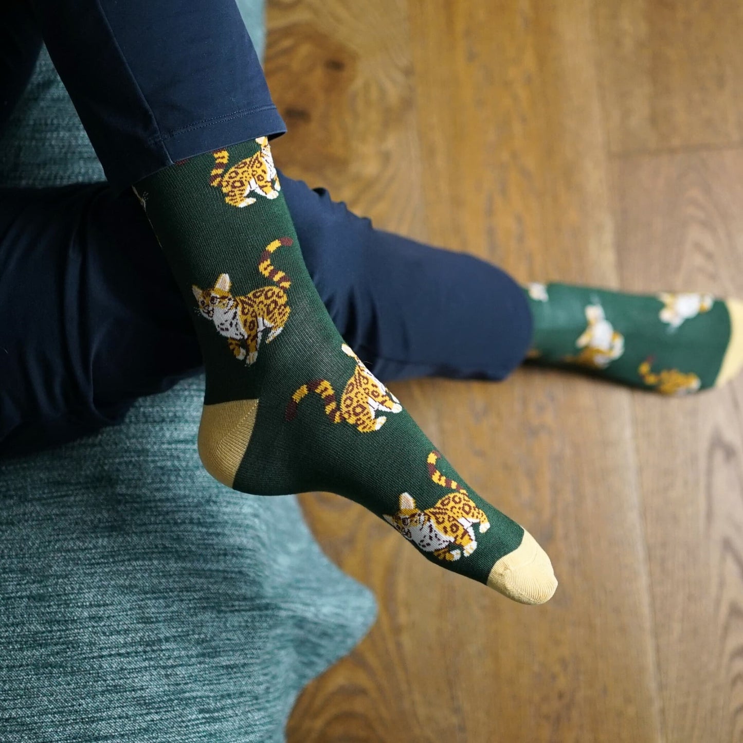 Green socks with leopard cat pattern worn by a person sitting on a wooden floor.