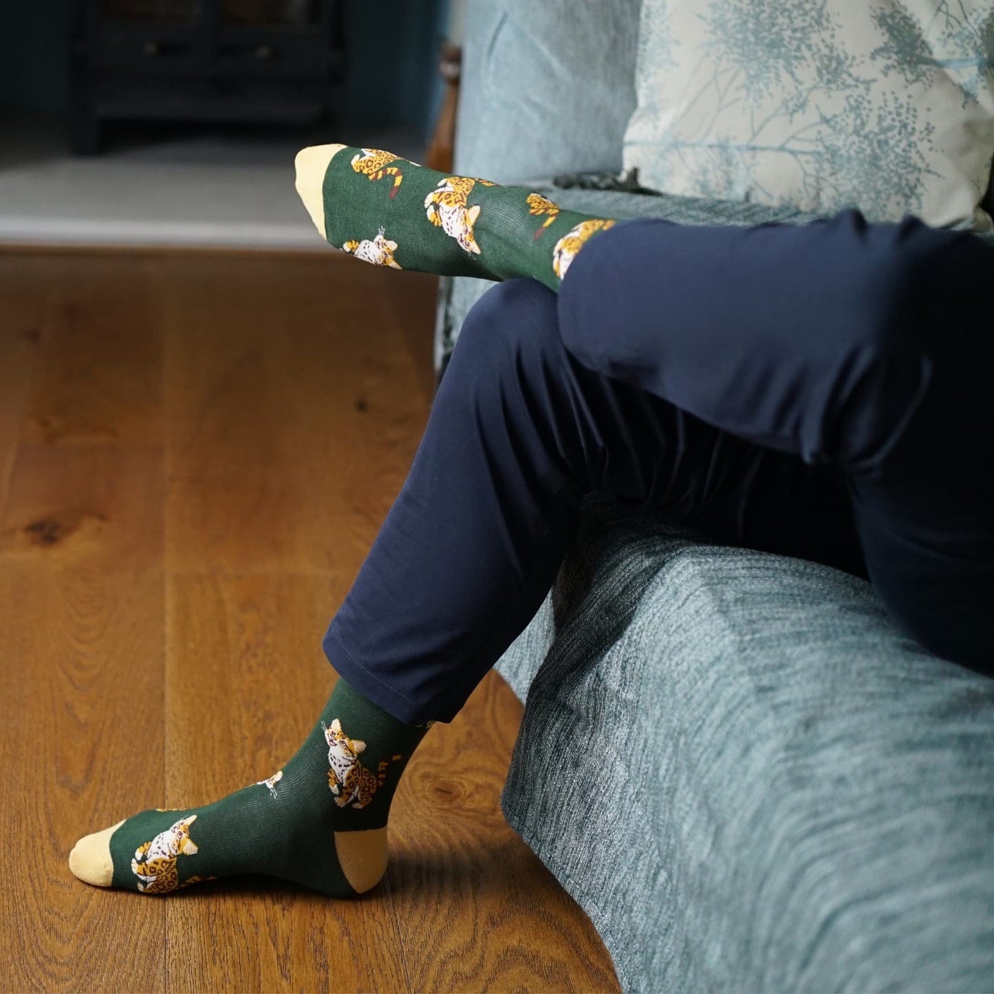 Green socks with leopard cat pattern worn by a person sitting on a wooden floor.