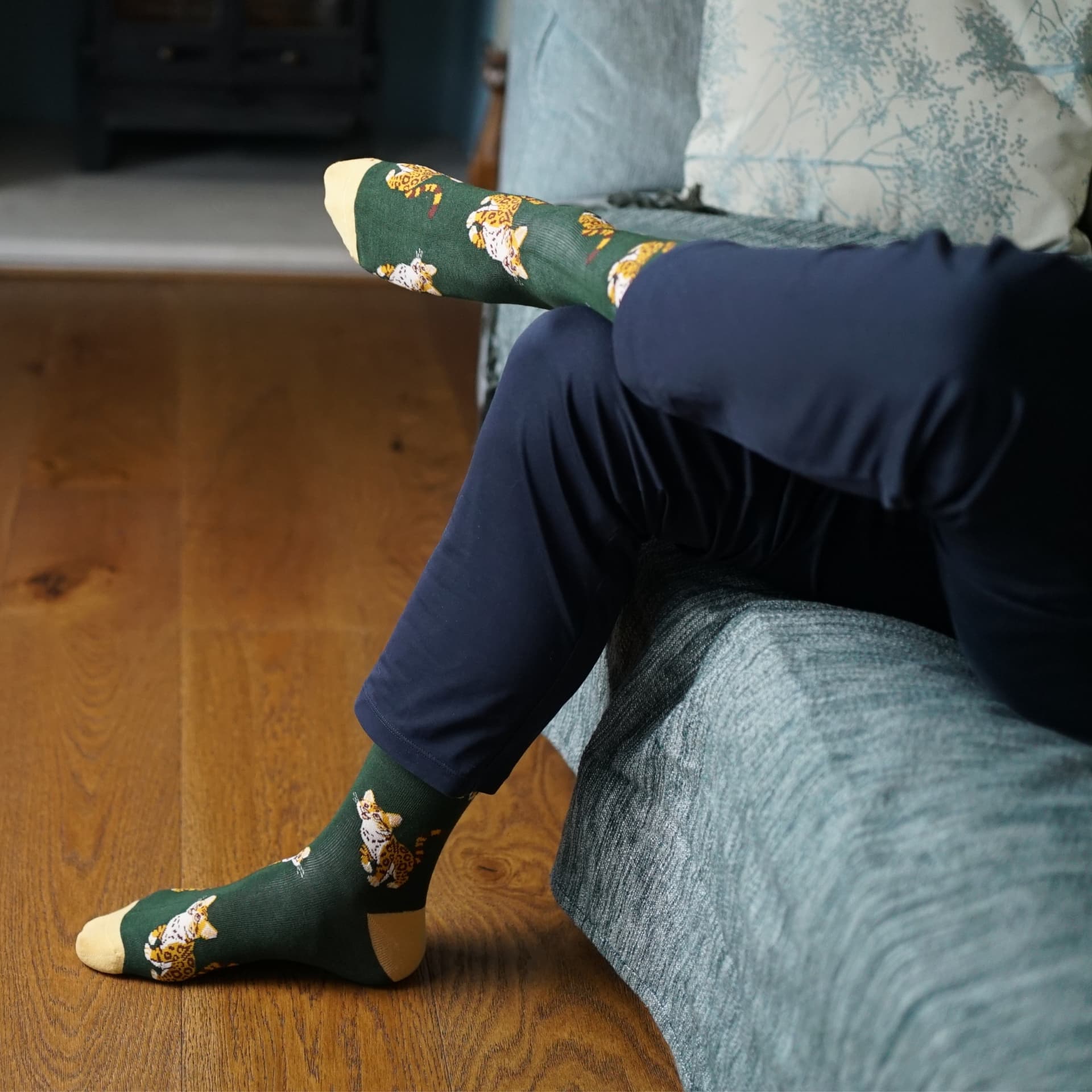 Green socks with leopard cat pattern worn by a person sitting on a wooden floor.