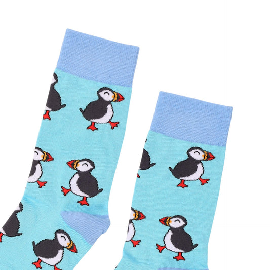 A pair of light blue crew socks with a repeating puffin pattern, featuring black and white bodies, red-orange beaks and feet, and light blue ribbed cuffs.