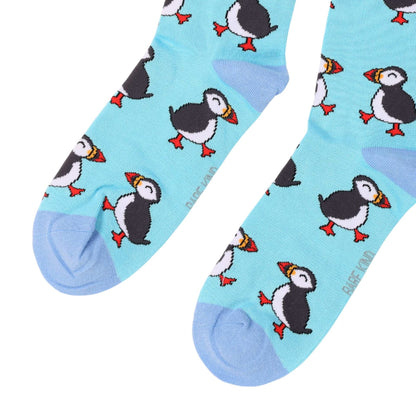 A pair of light blue crew socks with a repeating puffin pattern, featuring black, white, yellow, and orange puffins with light blue heels and toes, on a white background.