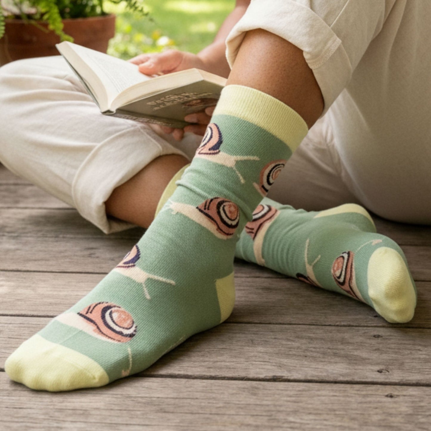 Person wearing green socks with snail design, sitting on a wooden deck reading a book.