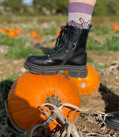 Person wearing black boots and a sock with a bat design, standing on a pumpkin in a field.
