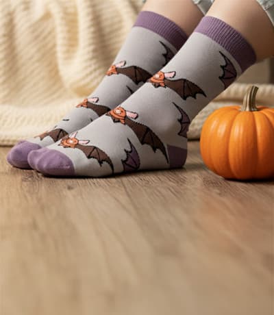 Purple socks with bat designs worn by a person, next to a small pumpkin on a wooden floor.