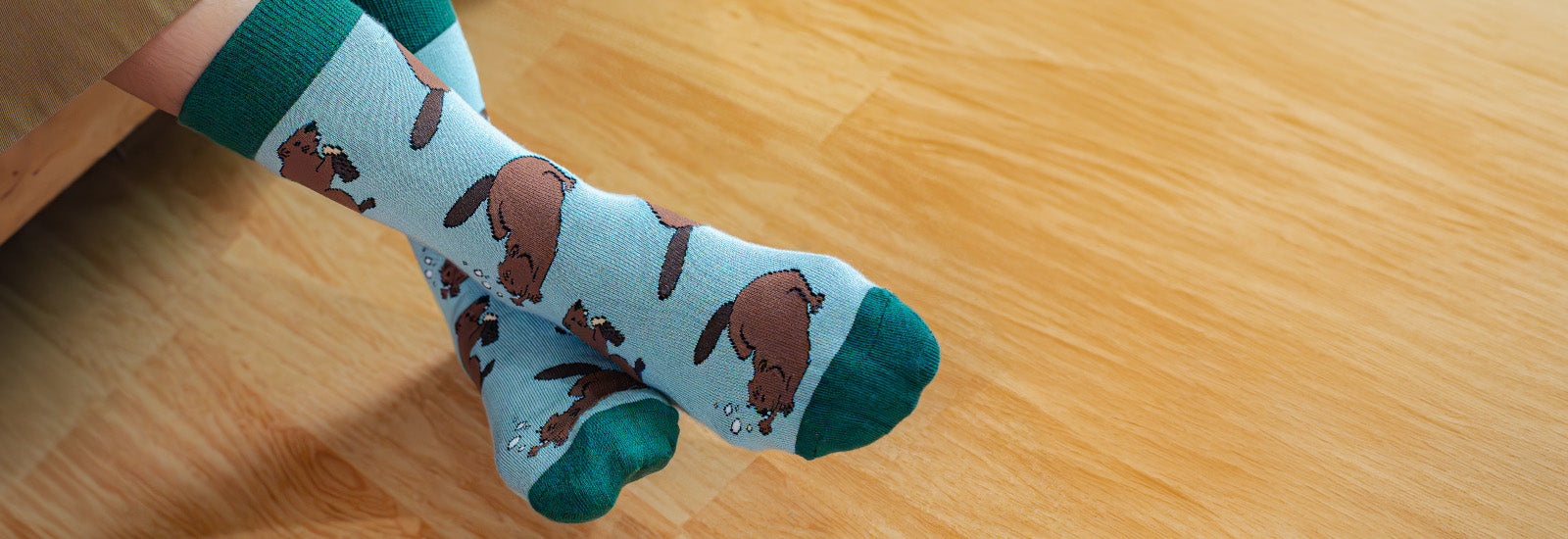 A pair of light blue crew socks with a brown beaver pattern and dark green toes and cuffs, worn on a person's feet on a light wood floor.