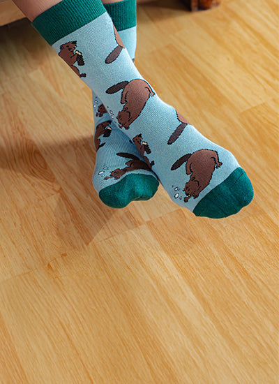 A pair of light blue crew socks with a brown beaver pattern and dark green toes and cuffs, worn on a person's feet on a light wood floor.