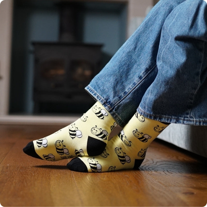 Person wearing yellow socks with black bee pattern and blue jeans on a wooden floor.