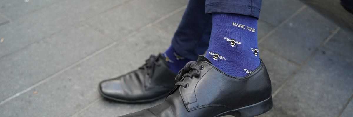 A close-up shot of a person's legs and feet, showing navy blue dress socks with a white and yellow pattern and the brand name 'BARE KIND' printed on them, paired with black leather dress shoes and dark trousers, set against a gray paved outdoor background.