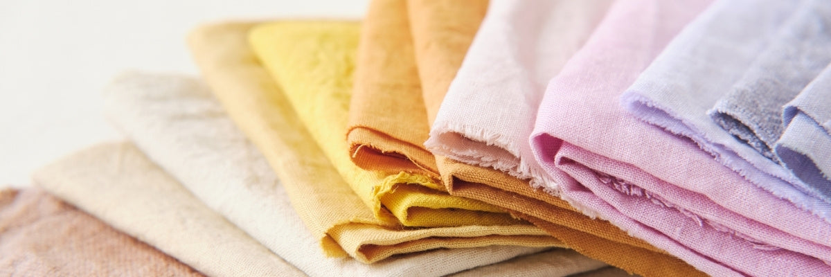 A stack of folded cotton fabric cloths in soft pastel and warm neutral tones, including beige, cream, yellow, peach, pink, and lavender, arranged diagonally on a white background.