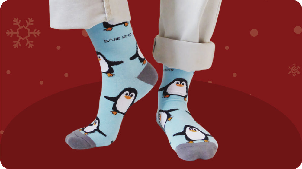 Light blue socks with penguin design worn by a person on a red background with snowflake patterns.