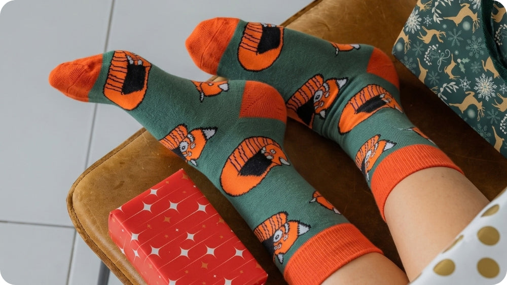 A pair of crew red panda socks, worn on feet resting on a brown leather couch next to a red gift box.