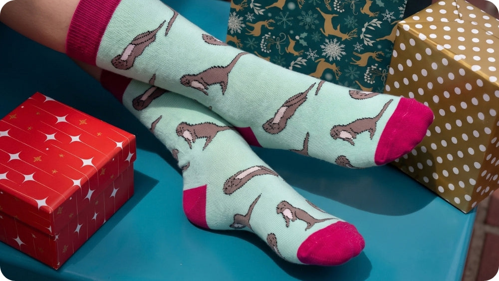 A pair of light mint otter socks with pink toes and heels, worn on feet resting on a blue surface next to wrapped gift boxes.