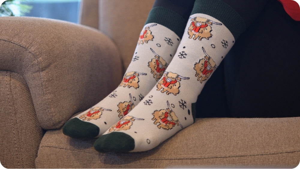 A pair of white christmas highland cow socks, dark green toes and cuffs, worn on a person's legs sitting on a beige couch.