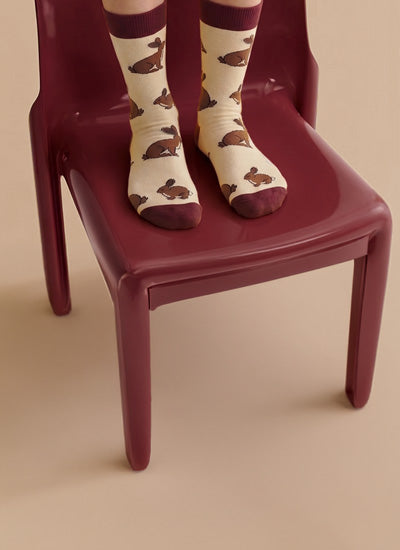 A person's feet wearing cream-colored socks with a brown hare pattern and dark burgundy cuffs, sitting on a dark burgundy plastic chair against a solid beige background.