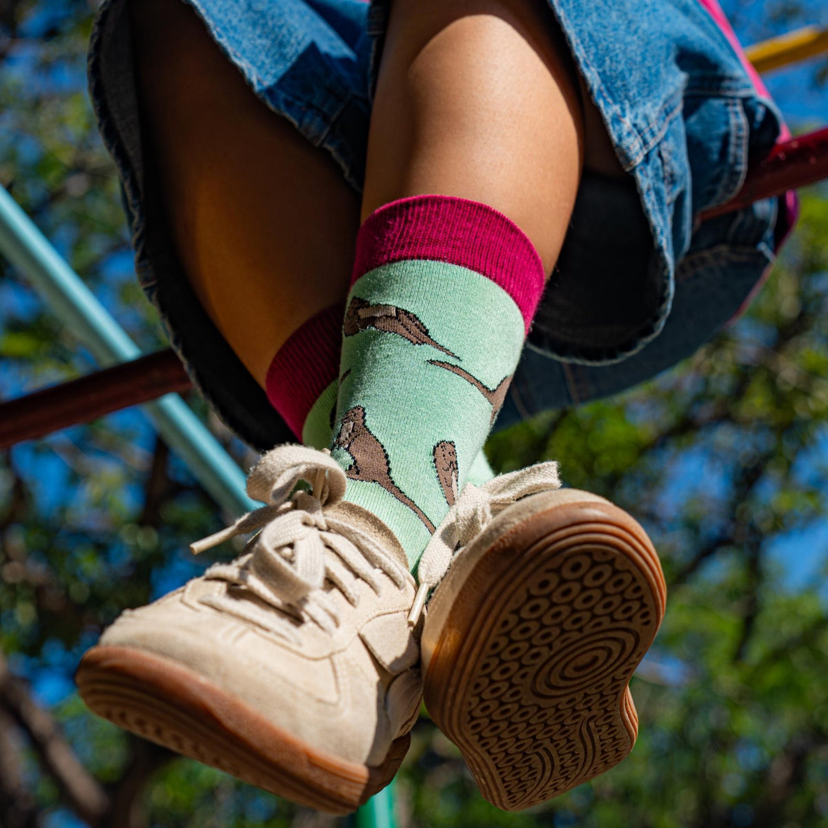 A close-up lifestyle shot of a person's legs hanging from playground bars, wearing light green socks with a brown otter print, bright pink cuffs, and beige sneakers.