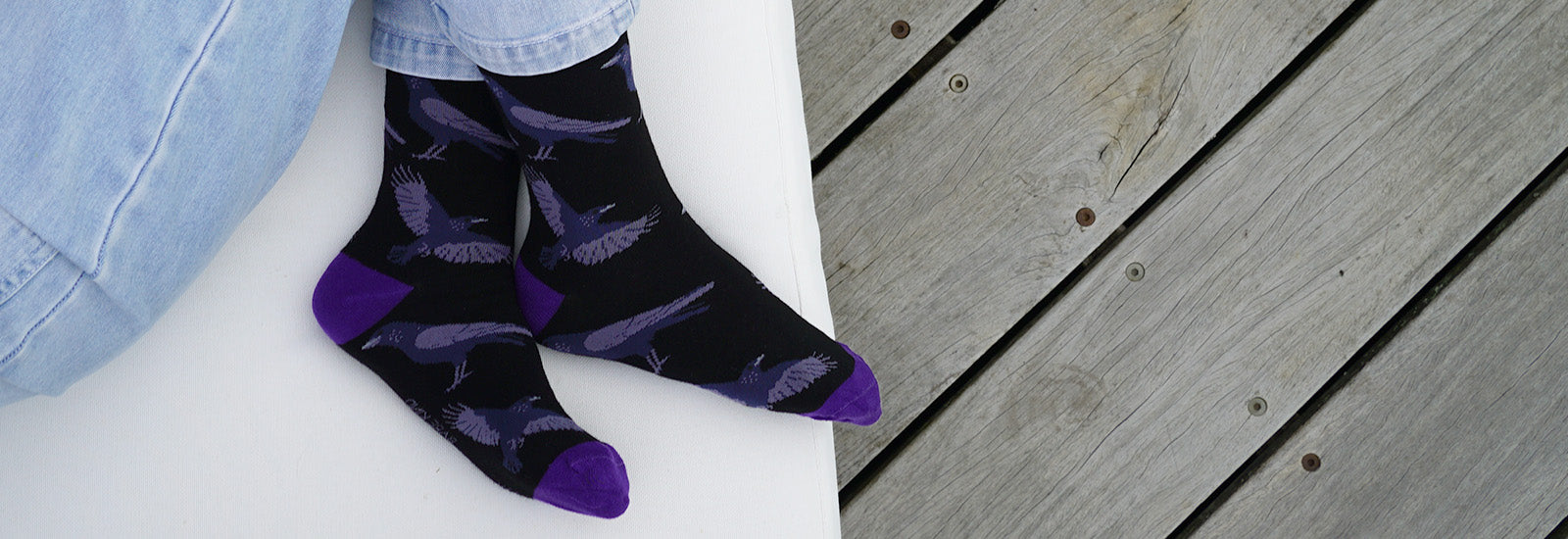 A close-up of a person's feet wearing black crew socks with a purple and gray raven pattern, sitting on a white bench with a gray wooden deck in the background.