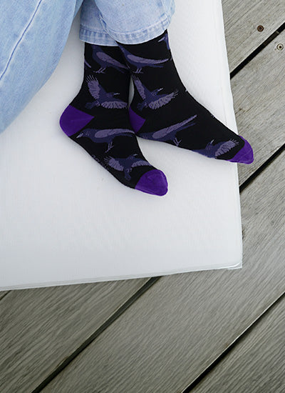 Black crew socks with a purple bird pattern and solid purple toes, heels, and soles, worn with light blue jeans on a white cushion over gray wooden deck planks.