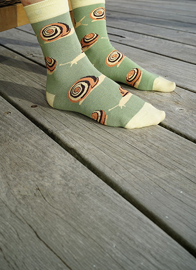 A pair of sage green crew socks with a pattern of orange, black, and cream snails, worn on feet standing on weathered gray wooden deck planks.