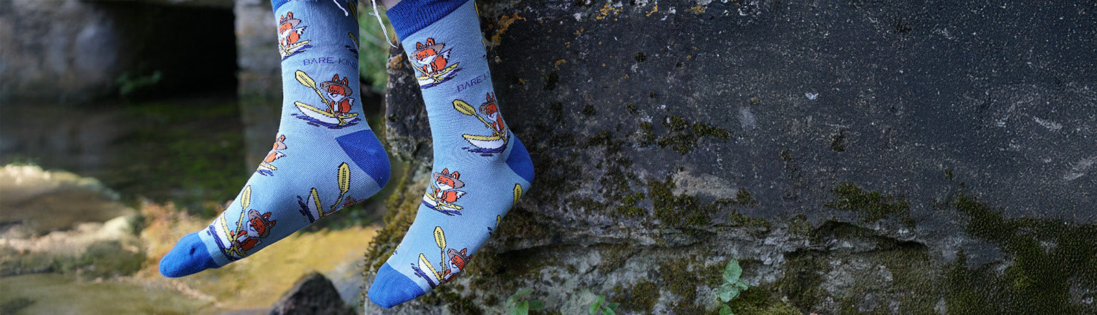 Person wearing blue sporty fox socks sitting on a stone by a stream.