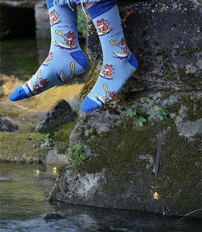 Person wearing blue sporty fox socks sitting on a stone by a stream.
