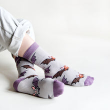 Model sits back with legs crossed in front, showing their left foot's sole and right foot's side, as they wear purple and grey bat bamboo socks